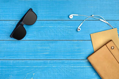 Black sunglasses, white earphones, brown craft paper sheet and leather folder on blue wooden desktop...