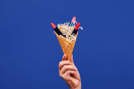 Hand of woman is holding a wafer cone filled with four open multicolored lipsticks against blue...