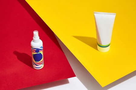 Colorful cosmetic tube and pump bottle with standing on red and yellow sheets of paper above white...