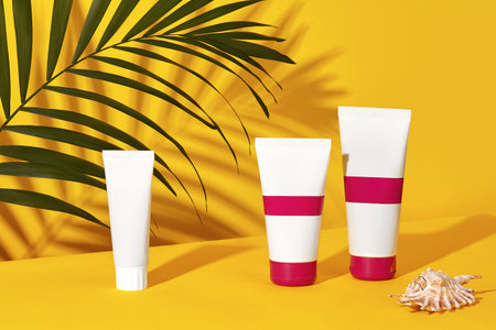 One white and two colorful cosmetic tubes next to a shell and green palm twig on yellow studio...
