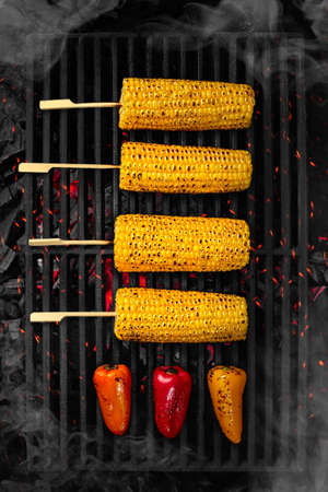 Ears of corn and bell peppers cooking on barbecue grate over smouldering coals
