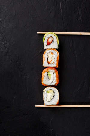 Four various sushi rolls with salmon, eel, shrimp, avocado lying on black textured surface between...