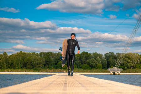 Athletic man in wetsuit carrying wakeboard, walking on pier after trainingの写真素材