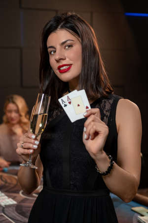 Happy young brunette with glass of champagne and set of winning cards standing near poker table