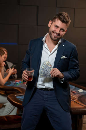 Bearded guy winning in poker, standing with glass of drink and banknotes in casinoの写真素材