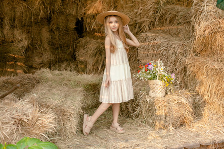Dreamy tween girl in light sundress and straw hat posing on hayloft with basket of wildflowersの写真素材