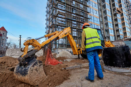 Professional builder in workwear and safety helmet standing near an excavator against modern...