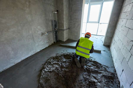 Unknown worker in protective clothes and orange safety helmet is making a cement floor screed in a...
