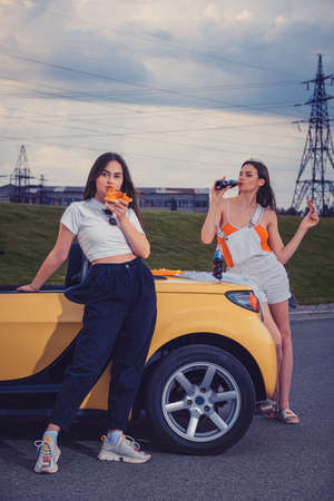 Attractive models in casual outfit eating pizza, enjoying soda water in glass bottles, posing near yellow car on parking lot. Full length, copy spaceの写真素材