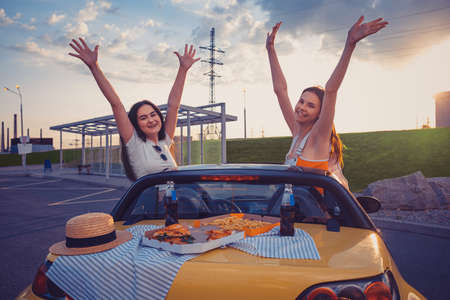 Ladies in casual outfit are smiling, raised up hands, posing in yellow car with french fries, pizza and soda on its trunk. Fast food. Copy spaceの写真素材