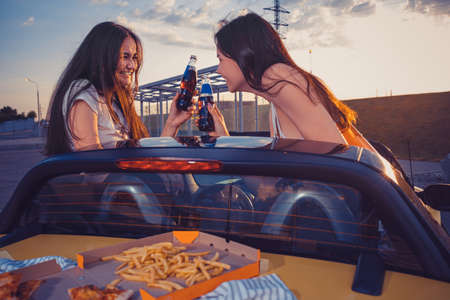 Happy women are smiling, cheering with soda in glass bottles, posing in yellow car with french fries...