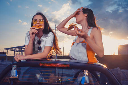 Happy models in casual outfit are eating pizza, posing in yellow car roadster with soda water in glass bottles on its trunk. Fast food. Copy spaceの写真素材