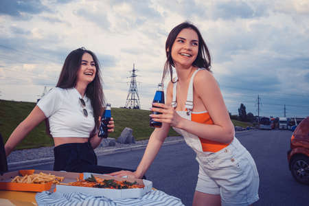Young ladies are smiling, enjoying soda in glass bottles, posing leaning on trunk of yellow car with...