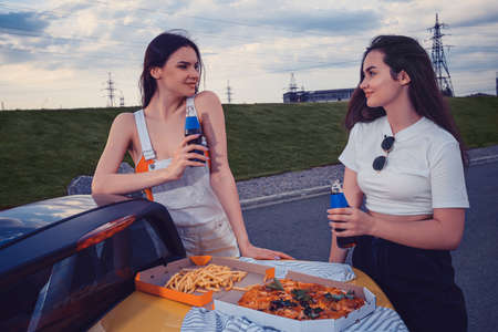 Young models are smiling, enjoying soda in glass bottles, posing leaning on trunk of yellow car with pizza and french fries on it. Copy spaceの写真素材