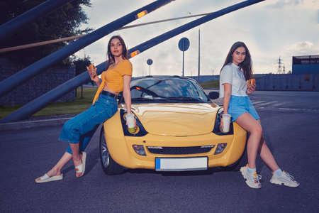 Gorgeous females in casual clothes are holding hamburgers and beverages in paper cups while sitting...