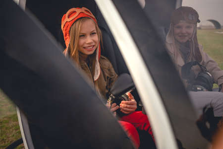 Smiling tween girl sitting in cockpit of landed helicopter near friend