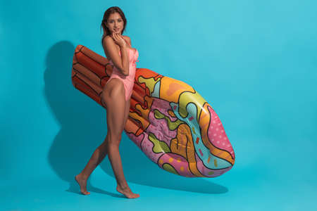 Smiling girl in bathing suit carrying swimming mattress on blue background