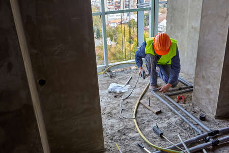Worker or builder in workwear and safety helmet is installing plastic pipes using modern tools in a...