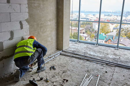 Builder in workwear and safety helmet is installing plastic pipes in flat of building under...