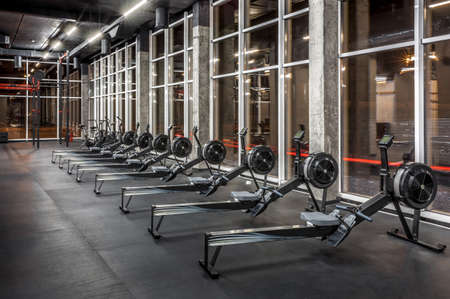 Modern rowing machines and exercise bikes lined near huge windows in spacious, empty gym interior. Equipment for physical training. Sport, fitnessの写真素材