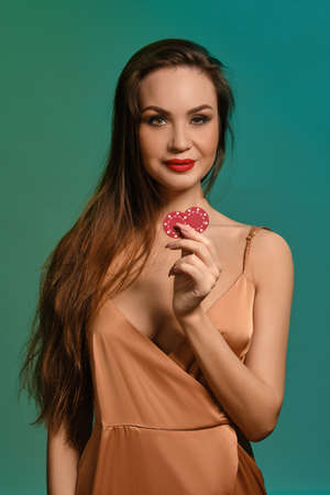 Brunette female in silk beige dress. She smiling, showing two red chips, posing against green background. Poker, casino. Close-upの写真素材