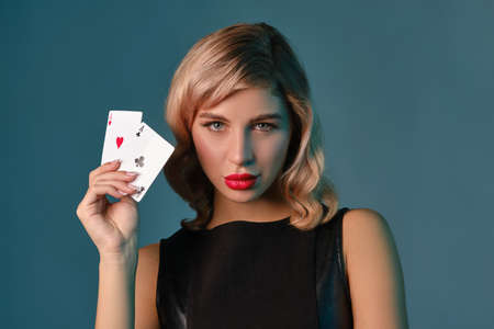 Blonde girl in black stylish dress showing two playing cards, posing against blue background. Gambling entertainment, poker, casino. Close-up.の写真素材