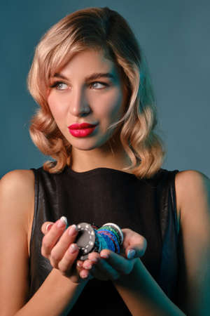 Blonde girl in black leather dress holding some colorful chips, posing against blue background. Gambling entertainment, poker, casino. Close-up.の写真素材