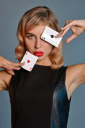 Blonde girl in black leather dress showing two playing cards, posing against gray background. Gambling entertainment, poker, casino. Close-up.の写真素材