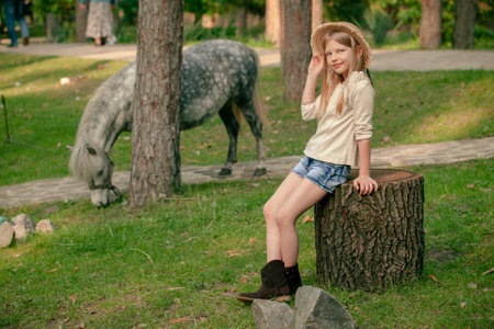 Cheerful tween girl sitting on stump in green park with gray pony grazing on grass in backgroundの写真素材