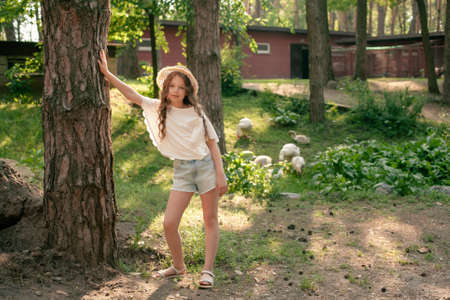 Full-length portrait of tween girl leaning of tree in courtyard of country house in summerの写真素材