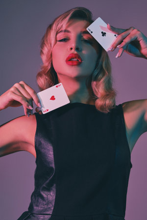 Blonde girl in black stylish dress showing two playing cards, posing against colorful background....