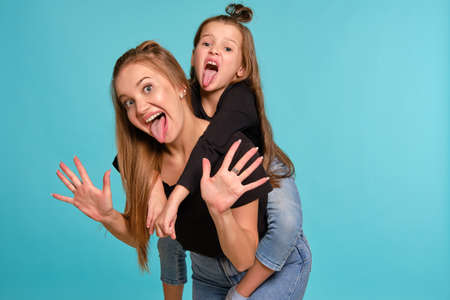 Mom and daughter with a funny hairstyles, dressed in black shirts and blue denim jeans are posing against a blue studio background. Close-up shot.の写真素材