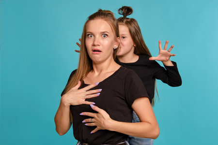 Mom and daughter with a funny hairstyles, dressed in black shirts and blue denim jeans are posing against a blue studio background. Close-up shot.の写真素材