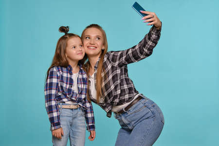 Mom and daughter dressed in checkered shirts and blue denim jeans are making selfie while posing...