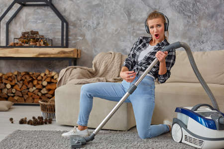 Full length portrait of cheerful young woman 20s listening to music via headphones and having fun with vacuum cleaner in the house.の写真素材