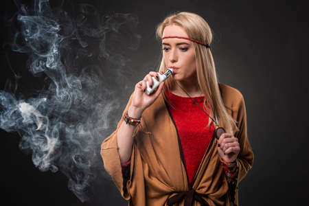 Portrait of sexy young hipster smoking vaping girl. The woman is dressed in the style of a boho. Vapor concept. Vaping e-Cigarette.の写真素材