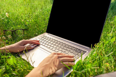 Close up hands on keyboard. Woman working on laptop pc computer with blank black empty screen to copy space in park on green grass sunshine lawn outdoors.の写真素材