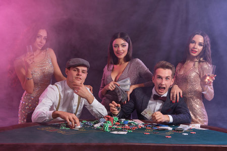 Friends playing poker at casino, at table with stacks of chips, money, cards on it. Celebrating win,...