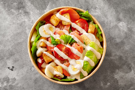 Paper bowl with Caesar salad of lettuce, tomatoes, quail eggs, croutons and smoked salmon on gray stone surfaceの写真素材