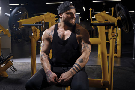 Tattooed, bearded athlete man in black sweatpants, vest and cap. He is looking thoughtful while sitting on an exercise machine at dark gym. Close upの写真素材