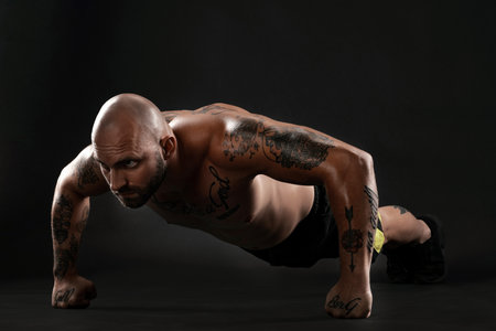 Athletic bald, tattooed man in black shorts and sneakers is posing against a black background....