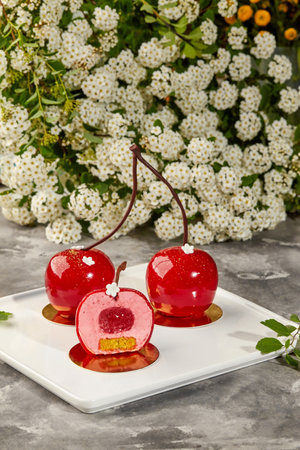 Cherry-shaped mousse cakes with mirror glaze on white plate displayed near white flowers