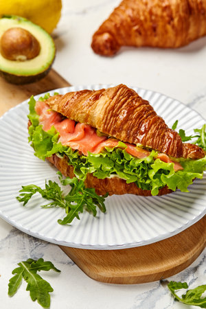 Delicious croissant sandwich with smoked salmon, avocado, and fresh greens being prepared on a wooden boardの写真素材