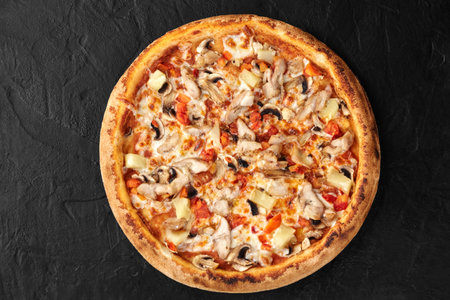 Delicious pizza with chicken, pineapple, and mushrooms resting on black surfaceの写真素材
