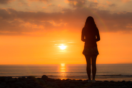 Young woman stands looking out to sea at the sunsetの写真素材