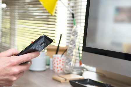Male hand holding a smart phone at desk with computerの写真素材