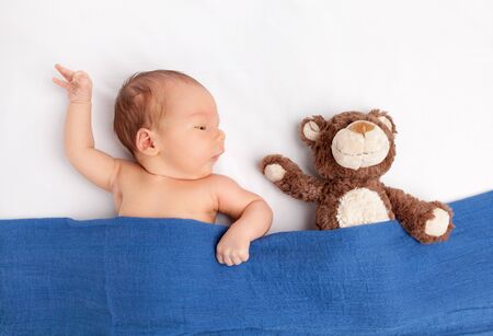 Cute newborn baby lying with a teddy bear on bed under a blanket