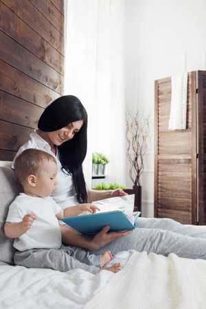 Child education. Happy mother with her toddler sitting on the bed and reading a book togetherの写真素材