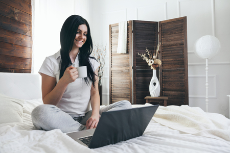 Relaxing on the bed. Beautiful young woman enjoying a cup of coffee and using her laptopの写真素材