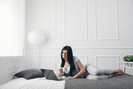 Relaxing on the bed. Beautiful young woman enjoying a cup of coffee and using her laptopの写真素材
